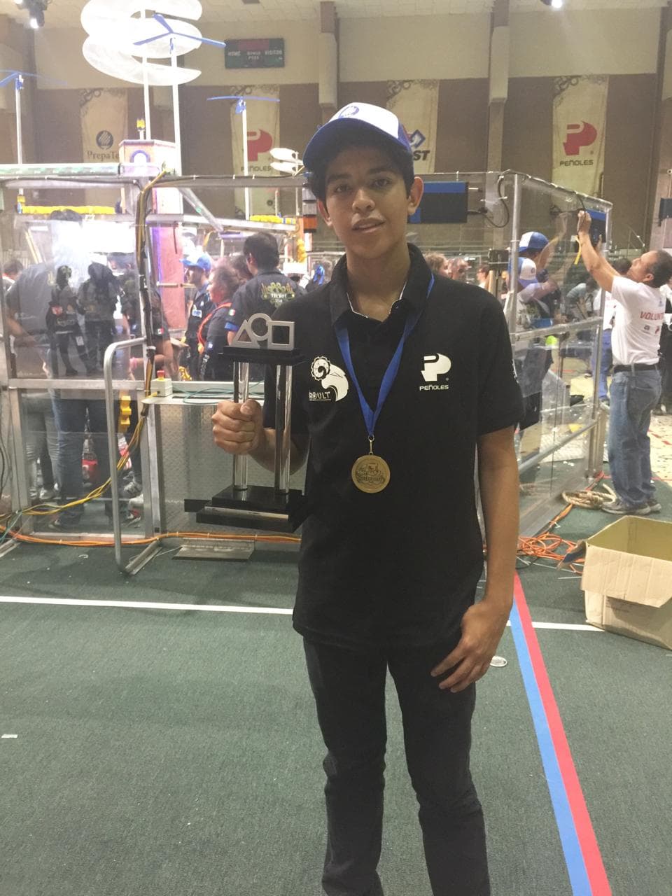 A participant wearing a medal holds a trophy beside a robotics competition field.