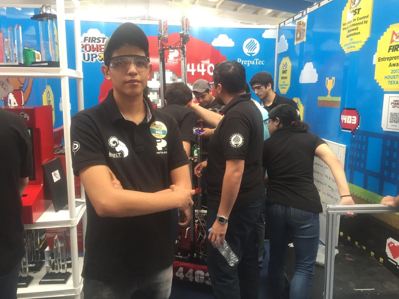 A participant in safety glasses stands in front of a group working on a robot in a blue team booth.