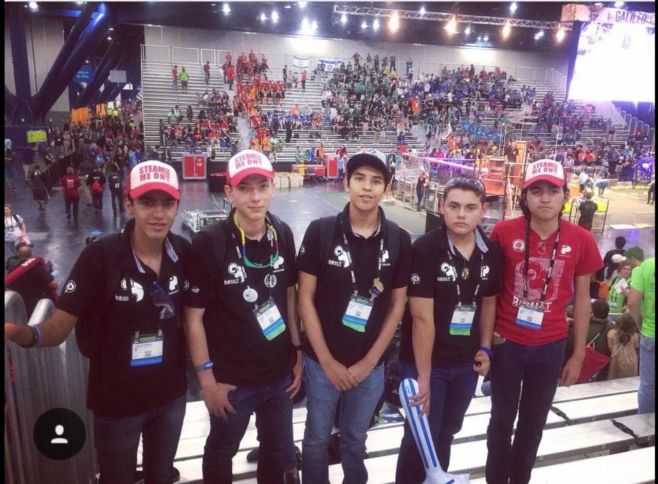 Five participants pose in the stands overlooking a busy robotics competition arena.