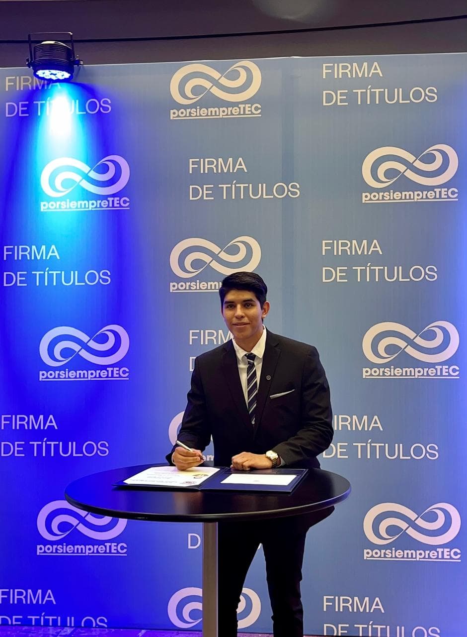A person in a suit holds a pen over a document at a table in front of a blue degree-signing backdrop.