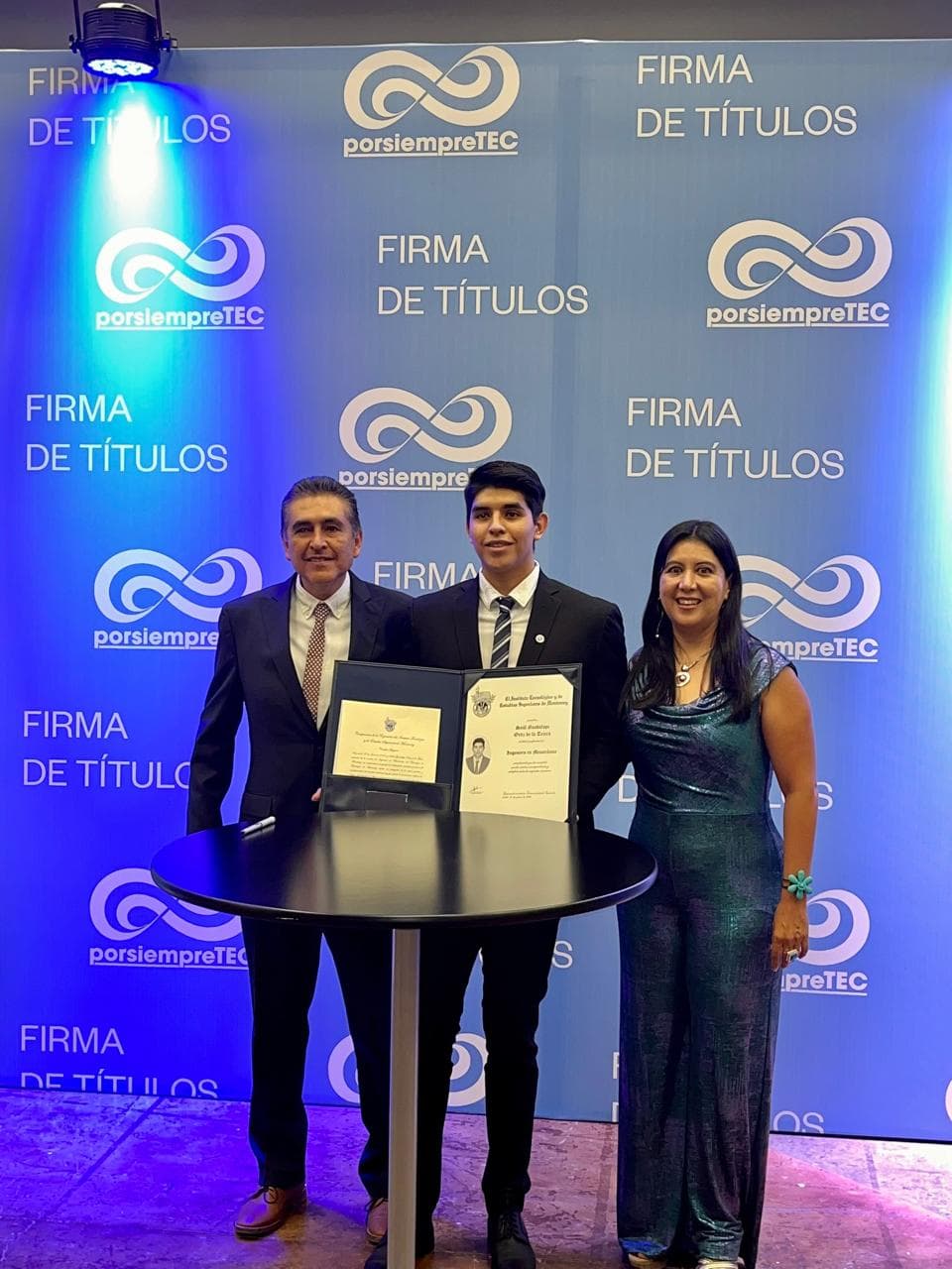 Three people pose behind an open diploma folder at a table in front of a blue degree-signing backdrop.