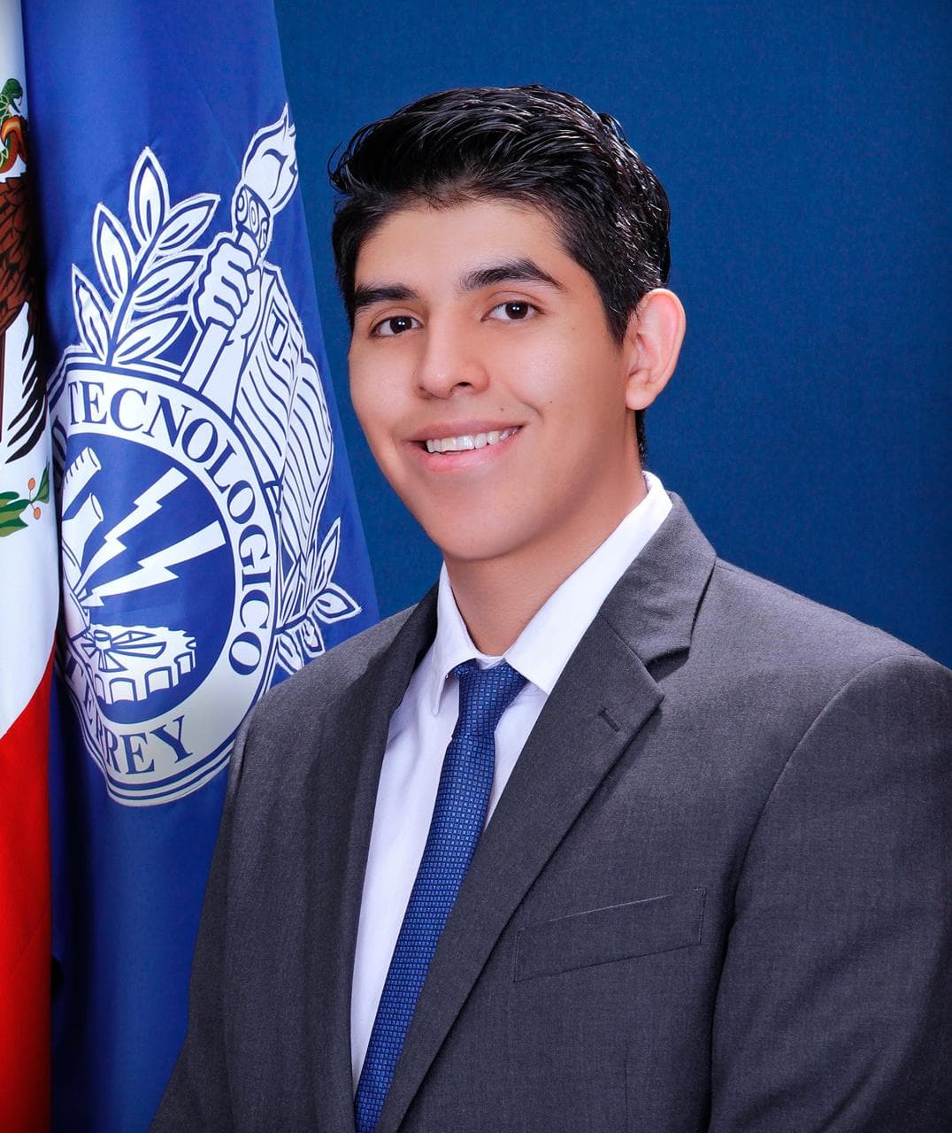 A formal portrait of a person in a grey suit and blue tie, with flags against a blue background.