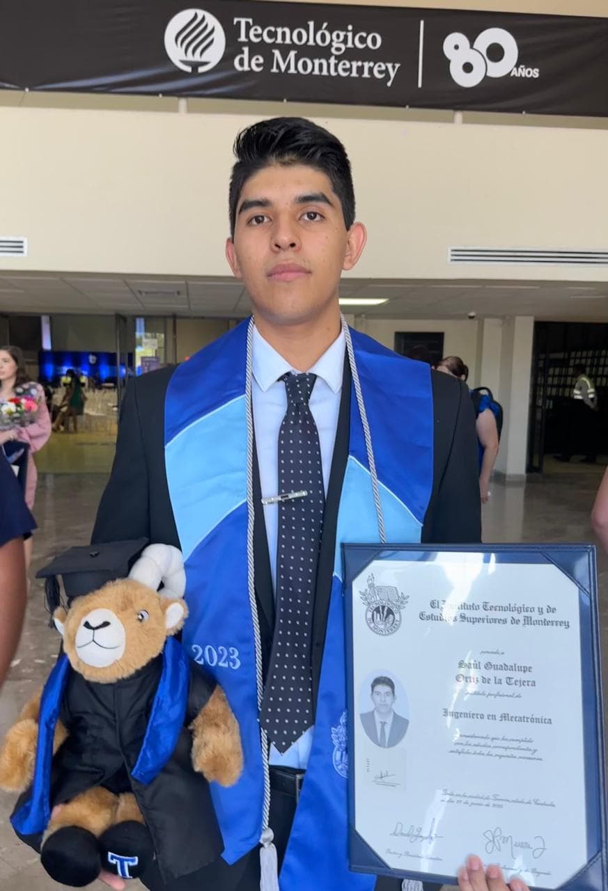 A graduate wearing a blue stole holds a framed diploma and a teddy bear in graduation clothing.